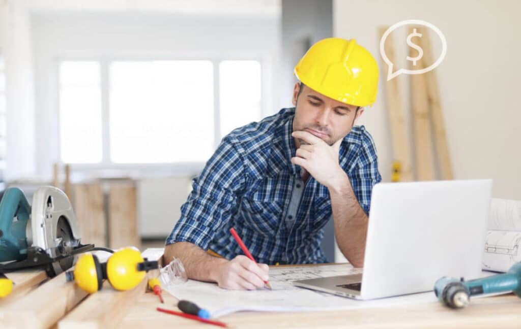 Contractor in a yellow hard hat at a laptop with a dollar sign speech bubble